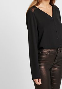 Czarna bluzka z długimi rękawami, dekoltem w kształcie V, zapięciem na guziki z przodu oraz koronkowym wykończeniem na ramionach. W połączeniu z błyszczącymi ciemnymi spodniami.