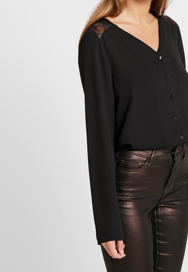 Czarna bluzka z długimi rękawami, dekoltem w kształcie V, zapięciem na guziki z przodu oraz koronkowym wykończeniem na ramionach. W połączeniu z błyszczącymi ciemnymi spodniami.