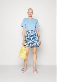 Blusa de mangas curtas azul claro combinada com uma saia floral azul. Os acessórios incluem uma bolsa de malha amarela e sandálias de plataforma cor lavanda.