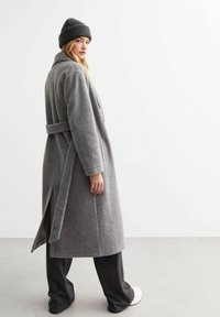 Grå ullrock med bälte i midjan, långa ärmar, sidofickor, parat med mörka byxor och en strukturerad svart stickad mössa.