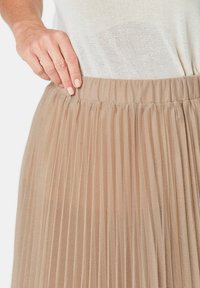Gelaagde beige tule rok met een elastische tailleband, die verticale strepen en een transparante textuur toont. Hand gepositioneerd bij de tailleband.
