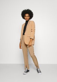 Blazer bege com um botão, gola alta preta, calças bege ajustadas com aberturas laterais e ténis altos pretos. Textura de tecido suave.