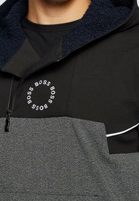 Sudadera con cremallera en negro y gris, con un alto cuello, tejido texturizado, impresión de logo en blanco y forro interior azul en contraste.
