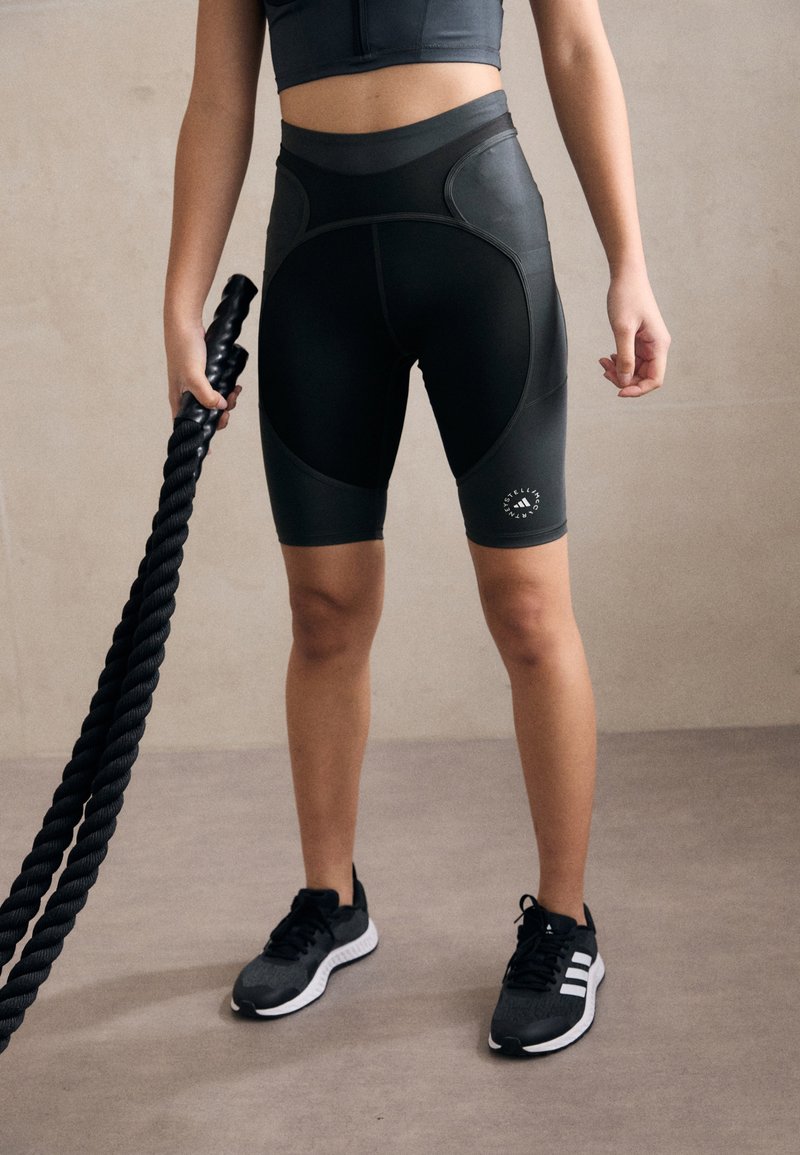 Black high-waisted athletic shorts with curved panel design, subtle logo detail, and smooth texture, paired with black running shoes.