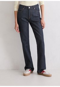 Dunkelblaue Denim-Jeans mit geradem Bein, ausgestattet mit kupferfarbenen Nähten, einem Knopfverschluss vorne und fünf Taschen. Kombiniert mit lässigen Sneakers.