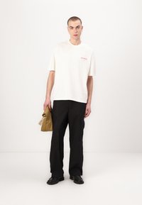 T-shirt blanche en coton avec logo rouge, associée à un pantalon cargo noir. Le modèle tient un sac olive plié, portant des bottes noires.