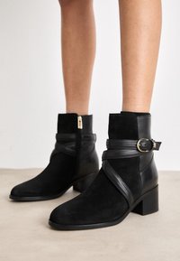 Stivaletti neri realizzati in suede e pelle, con tacco basso a blocco, zip laterale e una cintura decorativa con una fibbia dorata.