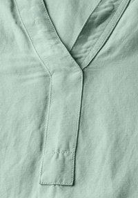 Helles grünes Leinengewebe mit einem V-Ausschnitt und sichtbaren Nahtdetails, das eine weiche Textur und einen leichten Glanz zeigt.