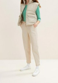 Väst i ljusbeige puffmaterial över en grön och vit randig långärmad skjorta, matchad med beiga byxor och vita sneakers. Släta texturer visas.