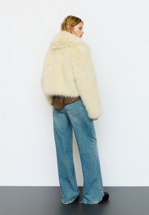 Giovane donna con capelli corti che indossa una giacca crema soffice, una camicia marrone a motivi, jeans larghi blu e scarpe nere, ritratta di profilo.