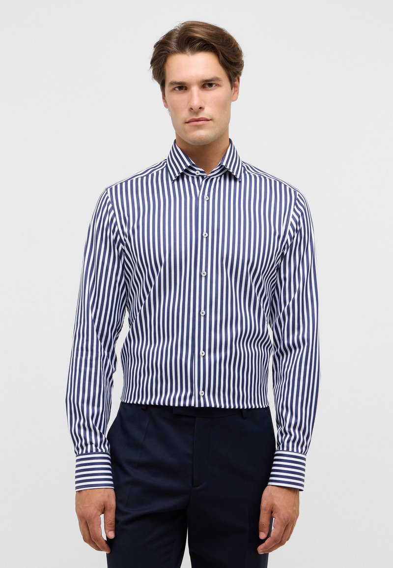 Man in een blauw-wit verticaal gestreept overhemd met knopen en donkere broek, staand tegen een lichte effen achtergrond.