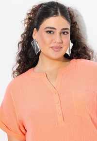 Camicia di colore corallo arancione con un tessuto texturizzato e stropicciato, maniche corte, colletto henley e tre bottoni. Presenta una tasca sul petto.