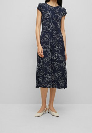 Femme portant une robe midi bleu marine à manches courtes avec des contours floraux blancs, assortie à des chaussures slingback blanches à petit talon.