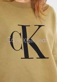La felpa verde oliva presenta un grande logo "CK" nero e la scritta "Calvin Klein Jeans" in bianco sotto. Realizzata in tessuto morbido e testurizzato.