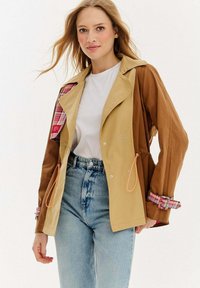 Svetlohnedá a hnedá bunda s kockovanou podšívkou, so striebornými doplnkami a oranžovými šnúrkami, s dlhými rukávmi a voľným strihom.