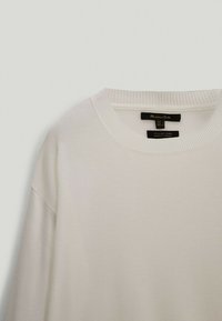 Vit tröja gjord av italiensk garn, med rund halsringning, ribbade ärmslut och en strukturerad stickning. Etikettuppgifterna är synliga.