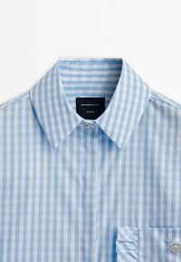 Camicia a righe blu chiaro e bianco con colletto classico, taschino sul petto e dettaglio decorativo sulla manica. Realizzata in tessuto intrecciato.