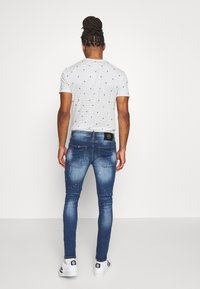 Man med flätat hår som bär en vit randig t-shirt, blå slitna slim-fit jeans och vita sneakers stående vänd mot en vit vägg.
