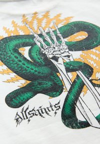 Grafiška marškinėlių nuotrauka su žalia gyvate, susipynusia su skeletinėmis rankomis, fone - balti marškinėliai su oranžiniais žaibų akcentais.