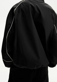 Zwarte nylonjas met witte bies langs de schouders en mouwen, voorzien van een ontspannen pasvorm en glad, gestructureerd materiaal.