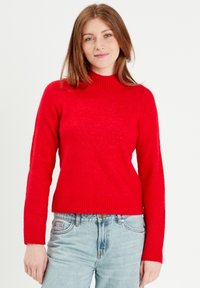 Pull en tricot rouge avec un col montant, des manches longues et un ourlet côtelé. Fabriqué en matériau doux, présenté avec un jean bleu.