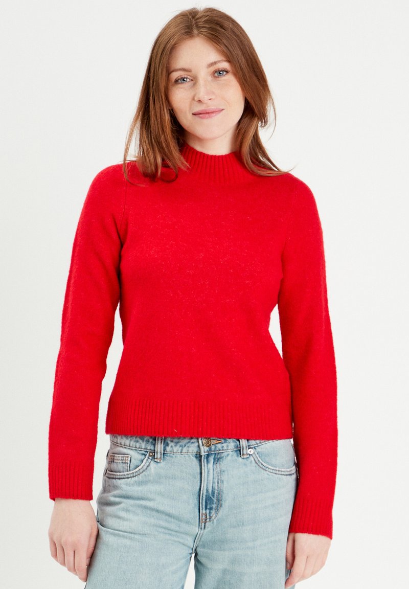 Pull en tricot rouge avec un col montant, des manches longues et un ourlet côtelé. Fabriqué en matériau doux, présenté avec un jean bleu.