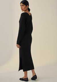 Abito lungo a costine nero con schiena a V, dettagli con spacco laterale e maniche lunghe. Indossato con scarpe nere flat.