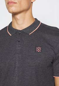 Homme portant un polo gris foncé avec col noir et finition rose, présentant un patch sur la poitrine avec "1962" et des lettres "T" croisées.