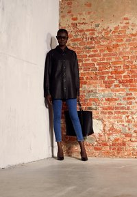 Camisa oversized preta de botões, jeans skinny azuis, sapatos de salto altos pretos e uma grande tote preta contra uma parede de tijolos texturizada.
