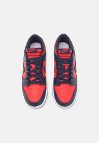Sneakers Nike con tomaia in pelle rossa, accenti blu navy e suole bianche; le caratteristiche includono la punta perforata e lacci spessi.