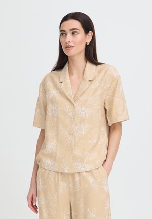 Donna con capelli scuri indossa una camicia beige a maniche corte con bottoni e pantaloni abbinati con motivi floreali bianchi, in piedi con una mano in tasca.