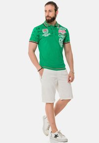 Grünes Polo-Shirt mit strukturiertem Muster und mehrfarbigen Akzenten, kombiniert mit weißen, hochgekrempelten Shorts und weißen Turnschuhen mit einem schwarzen Stern.