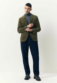 Hombre ajustando una chaqueta de pana verde sobre un suéter gris de cuello alto, vistiendo jeans azul oscuro y zapatos de cuero negros, de pie contra un fondo blanco.