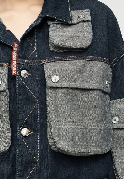 Τζιν μπουφάν με αντίθετες τσέπες σε ανοιχτό γκρι και ασημένια κουμπιά με σούστες, με ορατή κόκκινη ετικέτα DSQUARED2 κοντά στον γιακά.