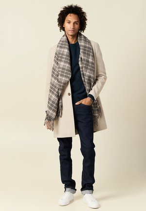 Cappotto di lana beige con un design colletto, maglione blu navy, jeans scuri e una grande sciarpa a scacchi bianca e nera. Sneakers bianche completano il look.