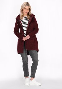 Burgunderfarbener langer Mantel aus strukturiertem Material, mit verstellbarer Kapuze und Taschen, kombiniert mit einem schwarz-weißen gestreiften Oberteil und grauen Jeans. Weiße Sneakers.