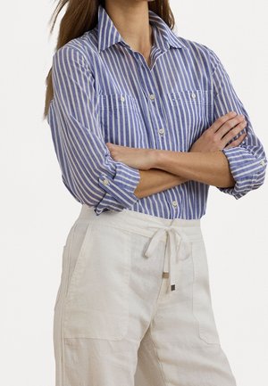 Frau mit langen Haaren trägt ein blau-weiß gestreiftes Hemd mit aufgekrempelten Ärmeln und weiße Hose mit Kordelzug, die Arme verschränkt.