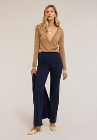Pull en métal doré à manches longues avec un col en V associé à un pantalon tailleur bleu marine. Modèle tenant une veste bleu marine, portant des chaussures à talons blocs jaune clair.