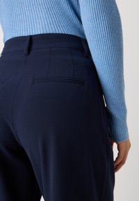 Marinefarbene Hosen aus glattem Stoff mit zwei Gesäßtaschen, kombiniert mit einem hellblauen gerippten Pullover.