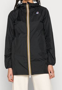 Schwarze Regenjacke mit Kapuze, ausgestattet mit einem Frontreißverschluss, der mit gelben, weißen und roten Streifen verziert ist. Elastische Bündchen und lockerer Schnitt.