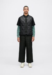 Chaleco negro acolchado sobre una camiseta negra de manga corta, combinado con pantalones anchos negros acolchados y zapatillas beige. Materiales texturizados con un ajuste relajado.