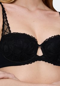 Reggiseno in pizzo nero con motivo floreale, dettagli a scalloped e un accento a forma di cuore al centro. Spalline regolabili e supporto con ferretto.
