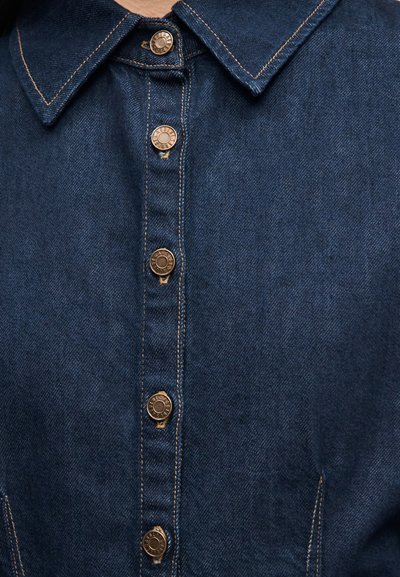 Camisa de mezclilla con textura azul, que presenta un cuello puntiagudo y botones de bronce en la parte delantera, acentuada por costuras amarillas.
