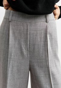 Grijze op maat gemaakte broek van een gestructureerde stof, met voorplooien en een platte tailleband, gepresenteerd tegen een donkere trui.