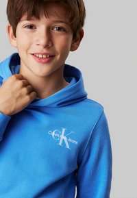 Sudadera azul de tejido suave que presenta un bolsillo tipo canguro y un logo blanco de Calvin Klein Jeans en el pecho. Textura suave, corte relajado.