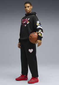 Sort hættejakke med farverige lapper og rød tekst, parret med sorte sweatpants med et hjerte-logo. Røde sneakers fuldender outfittet.