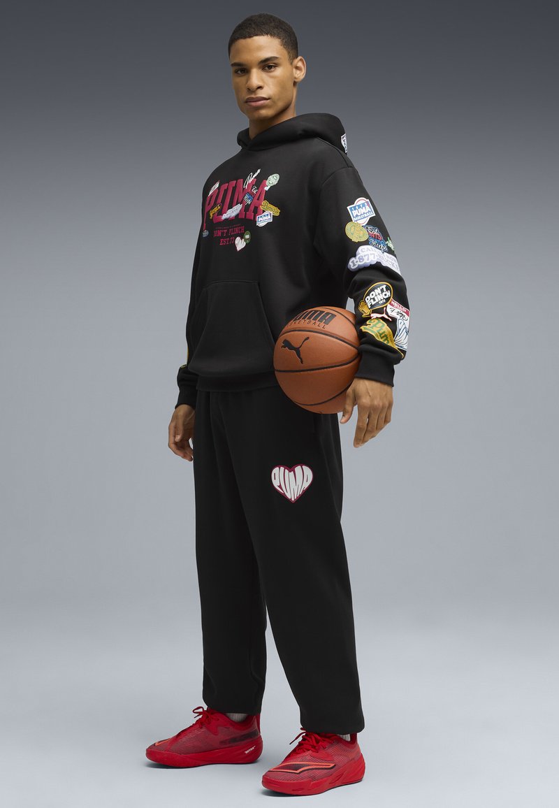 Sort hættejakke med farverige lapper og rød tekst, parret med sorte sweatpants med et hjerte-logo. Røde sneakers fuldender outfittet.