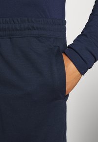 Persona vestida con pantalones azul marino y una camisa de manga larga, con la mano derecha metida en el bolsillo delantero de los pantalones.