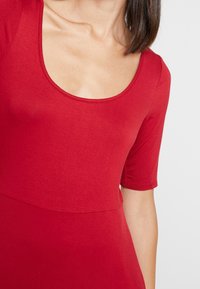 Punainen lyhythihainen mekko, jossa on avara kaula-aukko, valmistettu sileästä kankaasta. Sisältää vartalonmyötäisen yläosan ja levenevän hameen, jossa on minimaalinen ommel.