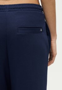 Κοντινό πλάνο ατόμου που φοράει σκούρο μπλε παντελόνι με ελαστική μέση, μικρή πίσω τσέπη με φερμουάρ και ακάλυπτη μέση.